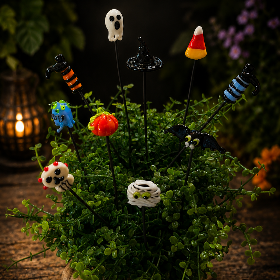 A set of Halloween-themed glass picket stakes with colorful sculpted toppers, displayed in a small green succulent arrangement. The toppers include a white ghost, a black spider, a blue monster, a red pumpkin, a black bat, a white spider, a striped orange/black witch, a blue/black spider, and a candy corn, set against a dark, atmospheric background with a warm lantern glow.