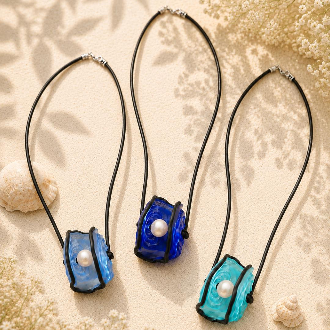 Three handmade glass pendant necklaces with black cords are arranged on a beige textured background with dried flowers and shells. The necklaces feature textured glass pendants in light blue, dark blue, and turquoise, each with a white faux pearl in the center.