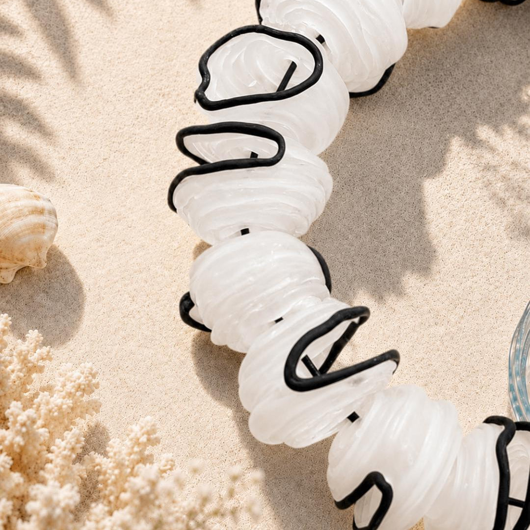 A close-up of a handmade beaded necklace with translucent white shell-shaped beads outlined in black, laid on sandy ground with seashells and coral nearby. Text in the upper left reads 'Handmade Details'.